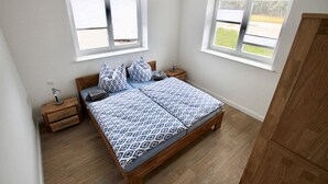Une chambre à coucher avec un lit en bois, deux tables de chevet et deux fenêtres équipées de stores.