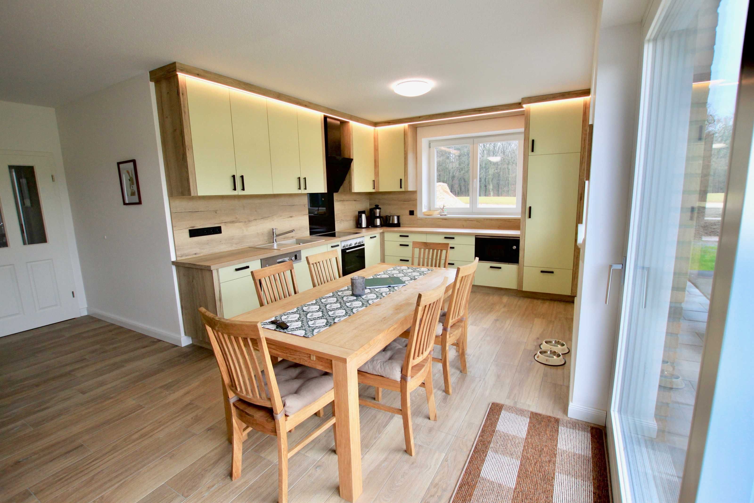 A modern kitchen with wooden cabinets, a dining table with chairs, and a large window providing natural light.