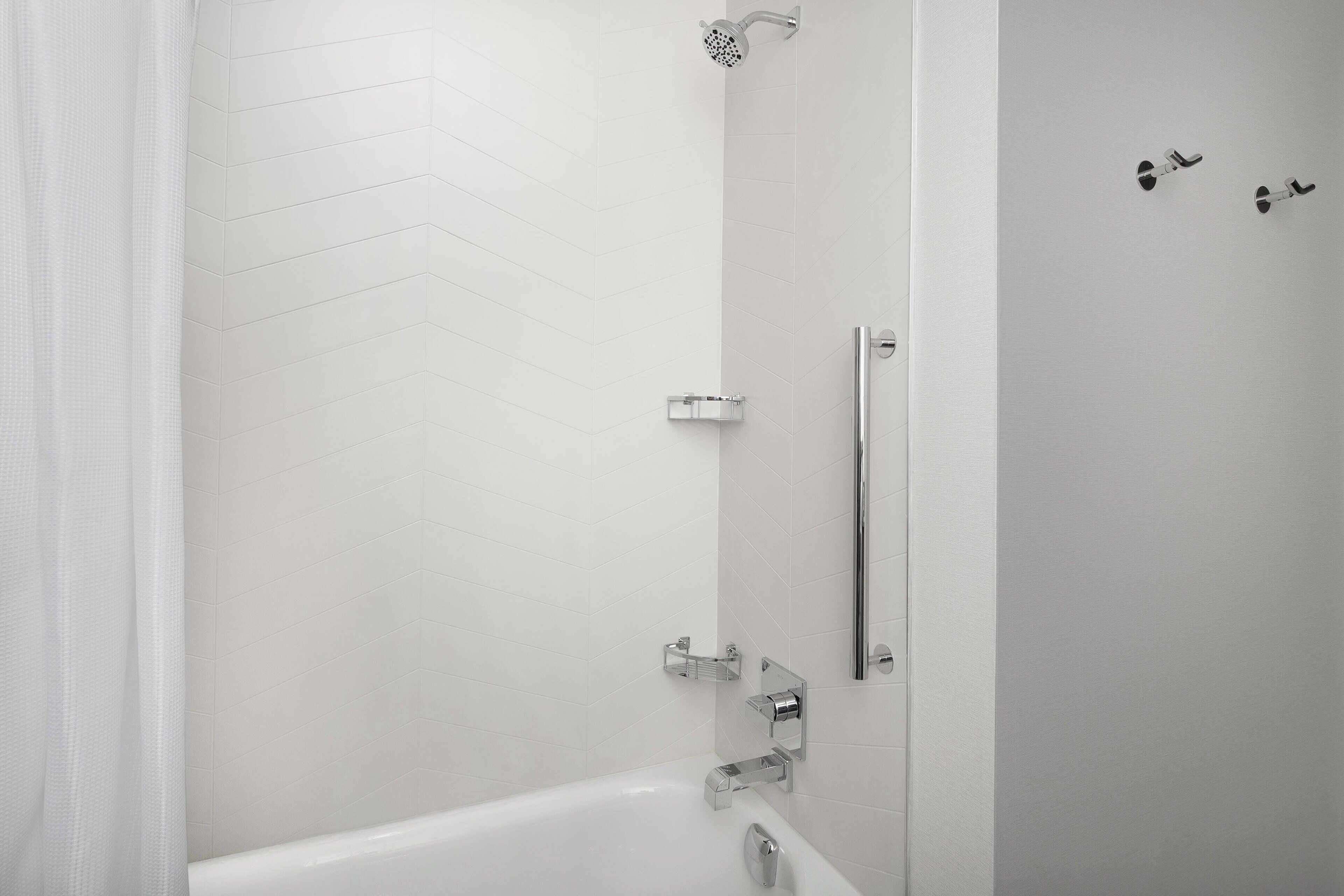 Ein modernes Badezimmer mit weiß gefliesten Wänden, einer Dusche mit Handbrause, einer Glasduschtrennwand und einer weißen Badewanne.