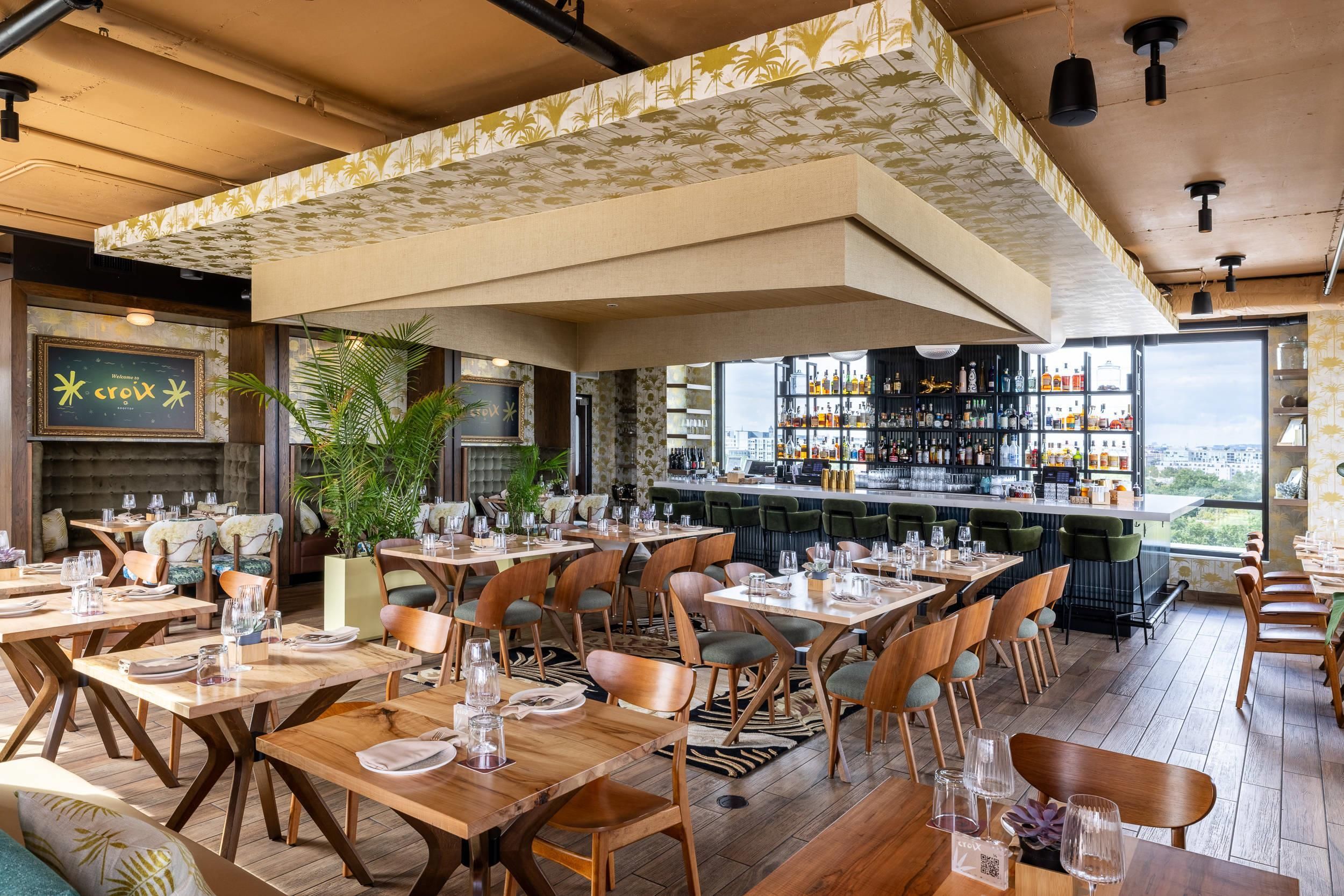Een restaurant met meerdere eettafels en stoelen, een bar met flessenkasten, en uitzicht door grote ramen.