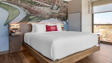 A modern hotel room with a large bed, a cityscape mural on the wall, a nightstand with a lamp, and a large window offering a view of the city.