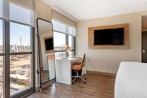Habitación de hotel con un ventanal grande, un escritorio con silla, una cama y un televisor montado.