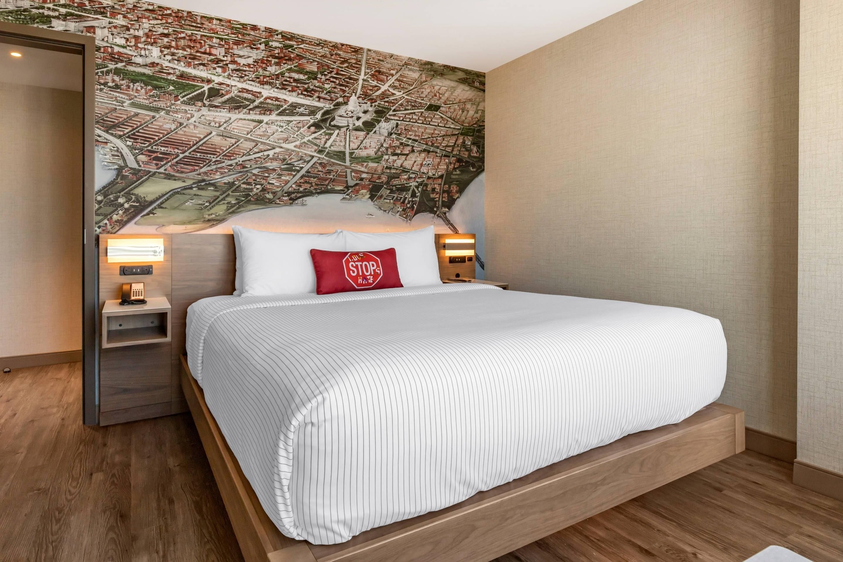 A modern hotel room with a large cityscape mural on the wall, a bed with a white striped bedspread, a red pillow with a stop sign, and bedside lamps.