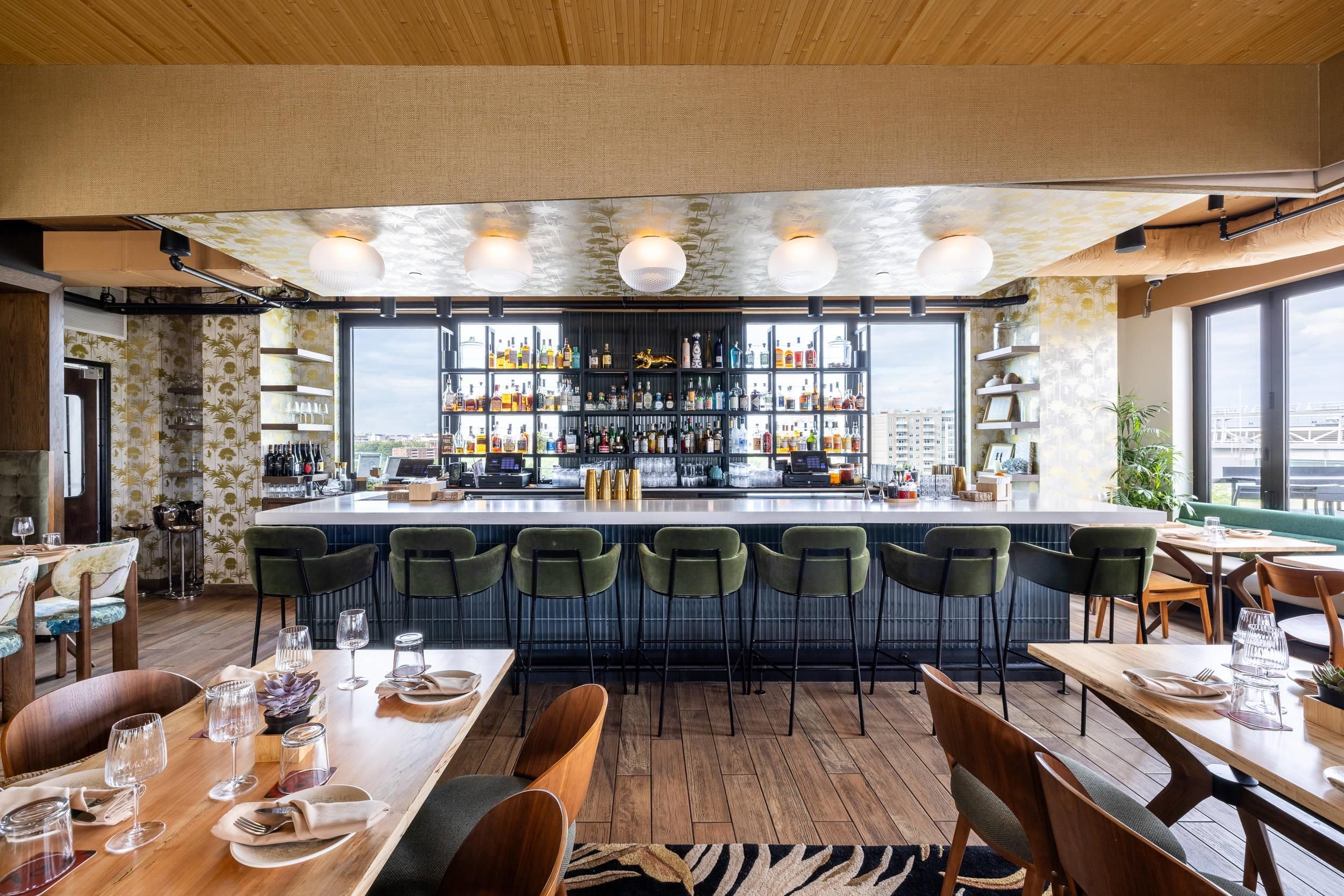 A bar area with a long counter, multiple shelves of bottles, and a view of the city through large windows.
