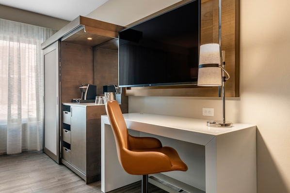 Ein modernes Hotelzimmer mit einem weißen Schreibtisch, einem braunen Ledersessel, einem flachbildfernseher an der Wand und einer Lampe auf dem Schreibtisch.