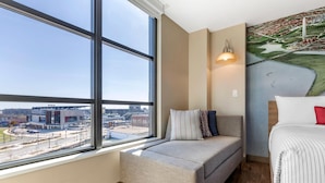 A modern hotel room with a large window offering a city view, a cozy seating area, a bed with striped bedding, and a wall-mounted lamp. - Cambria Hotel Washington D.C. Navy Yard Riverfront (Washington)