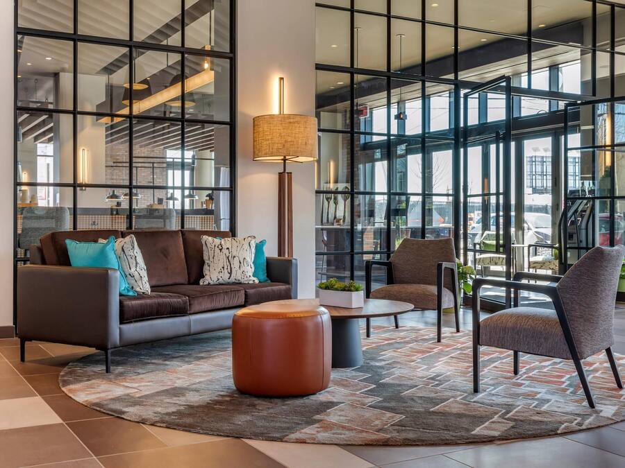 A modern lobby with a sofa, ottoman, and chairs, large windows, and a lamp.