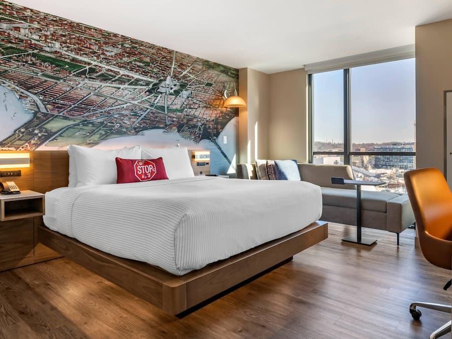 A modern hotel room with a large bed, a cityscape mural, a view through large windows, and a desk with a chair.