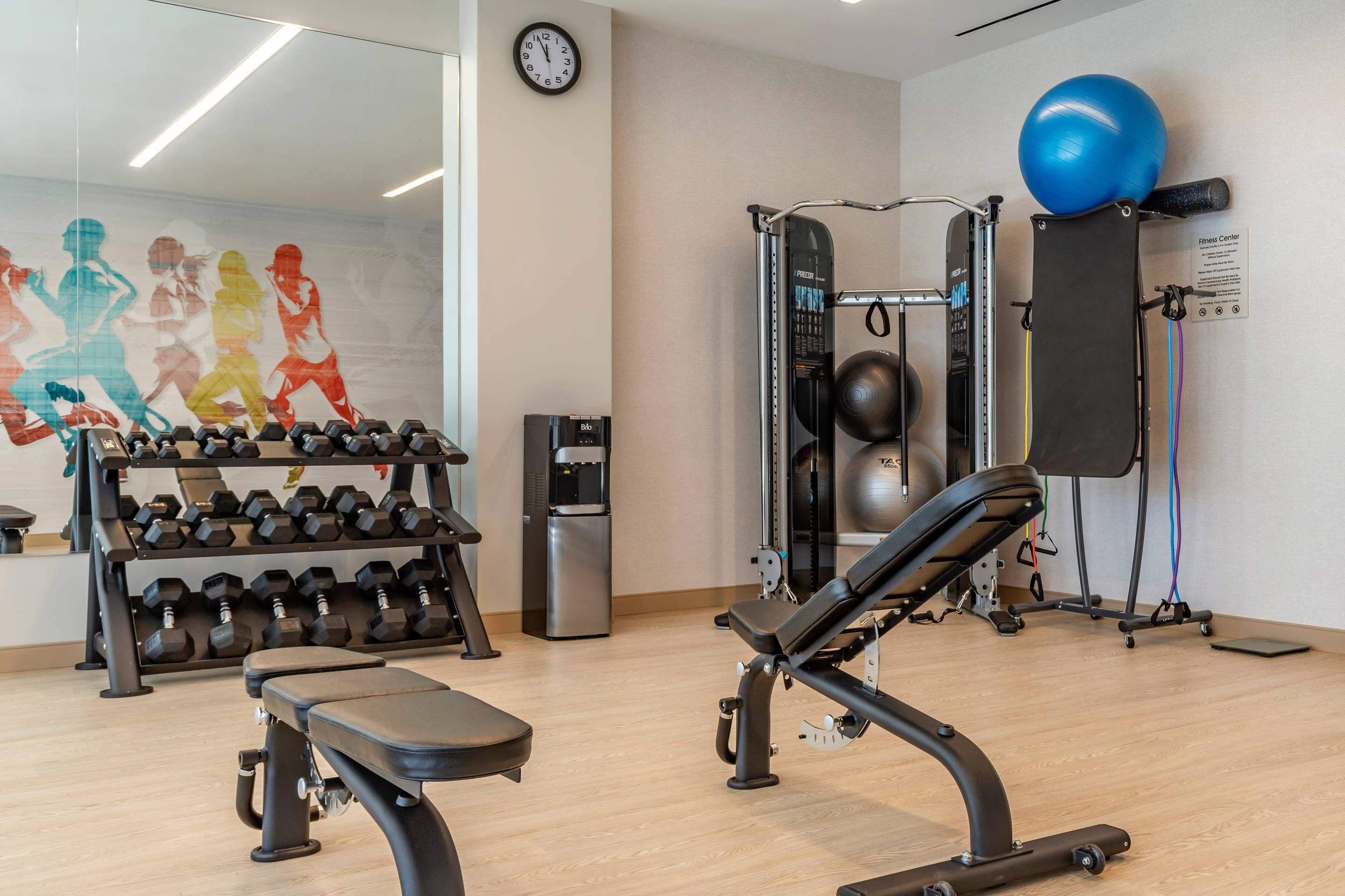 Ein Fitnessraum mit Gewichte, einer Bank, einer Wanduhr und einem Wasserspender.