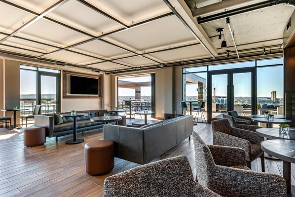 Un moderno espacio lounge con sofá, sillas y mesas, ventanas amplias que ofrecen vistas a la ciudad y un televisor de pantalla plana montado en la pared.