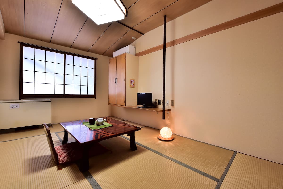 A traditional Japanese room with tatami flooring, a low wooden table, a tatami mat bench, a sliding door, a shelf with a television, and a hanging light fixture.