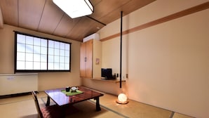 A traditional Japanese room with tatami flooring, a low wooden table, a tatami mat bench, a sliding door, a shelf with a television, and a hanging light fixture. - Guest House Takenoya (Omachi)