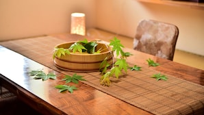 A wooden dining table with a woven placemat, a bowl with green leaves, and a candle in the background. - Guest House Takenoya (Omachi)
