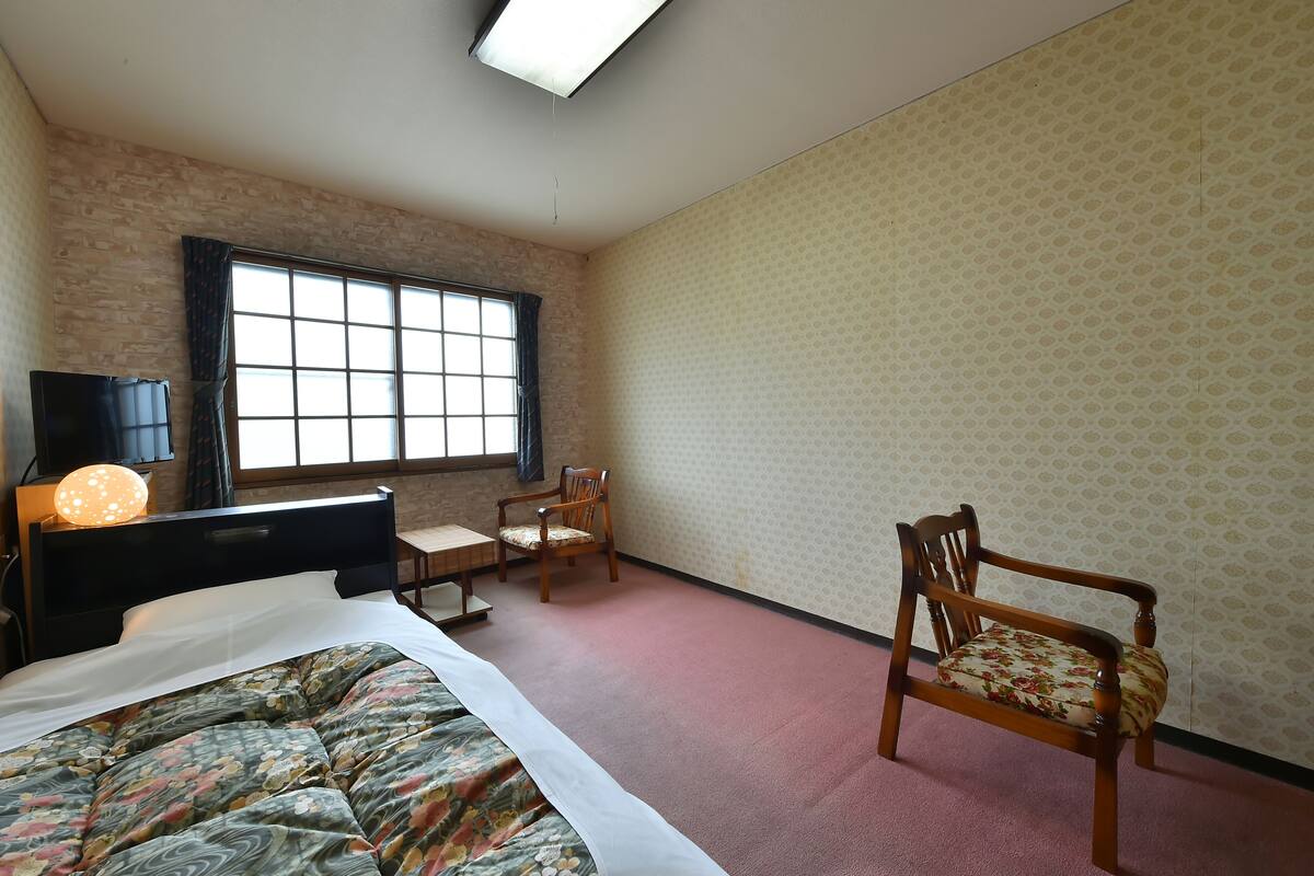 A bedroom with a bed, a desk, a chair, a TV, and a window.