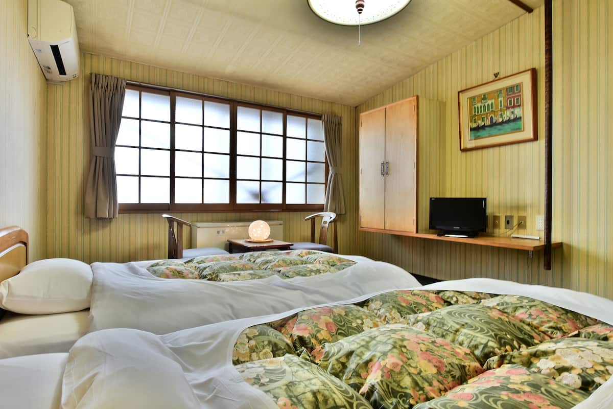A hotel room with two beds, a television on a shelf, a window with curtains, and a painting on the wall.
