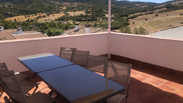 Un balcon avec une table et des chaises, donnant sur un paysage vallonné parsemé d’arbres et de bâtiments.