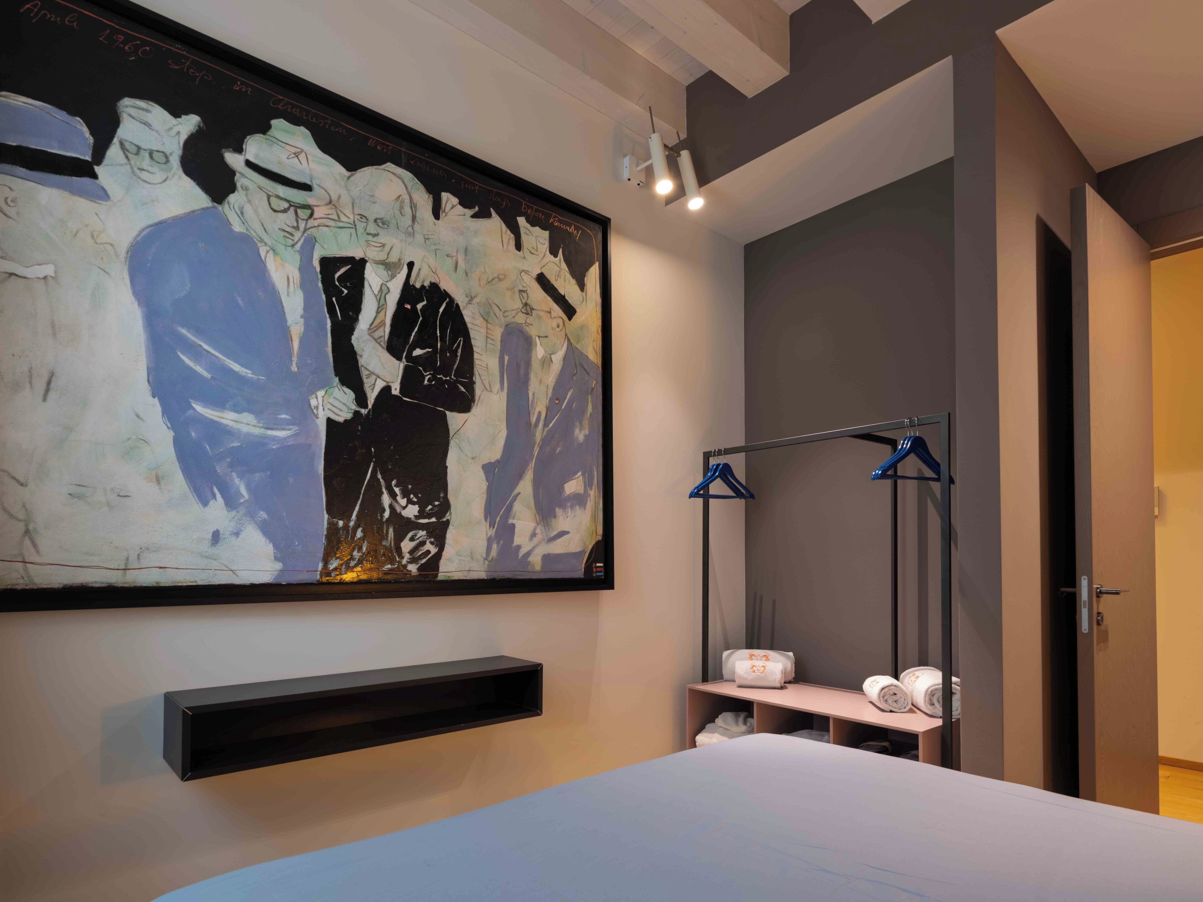 A modern hotel room with a large abstract painting on the wall, a bed with a white comforter, a small bedside table with a lamp, and a wardrobe with a mirror.