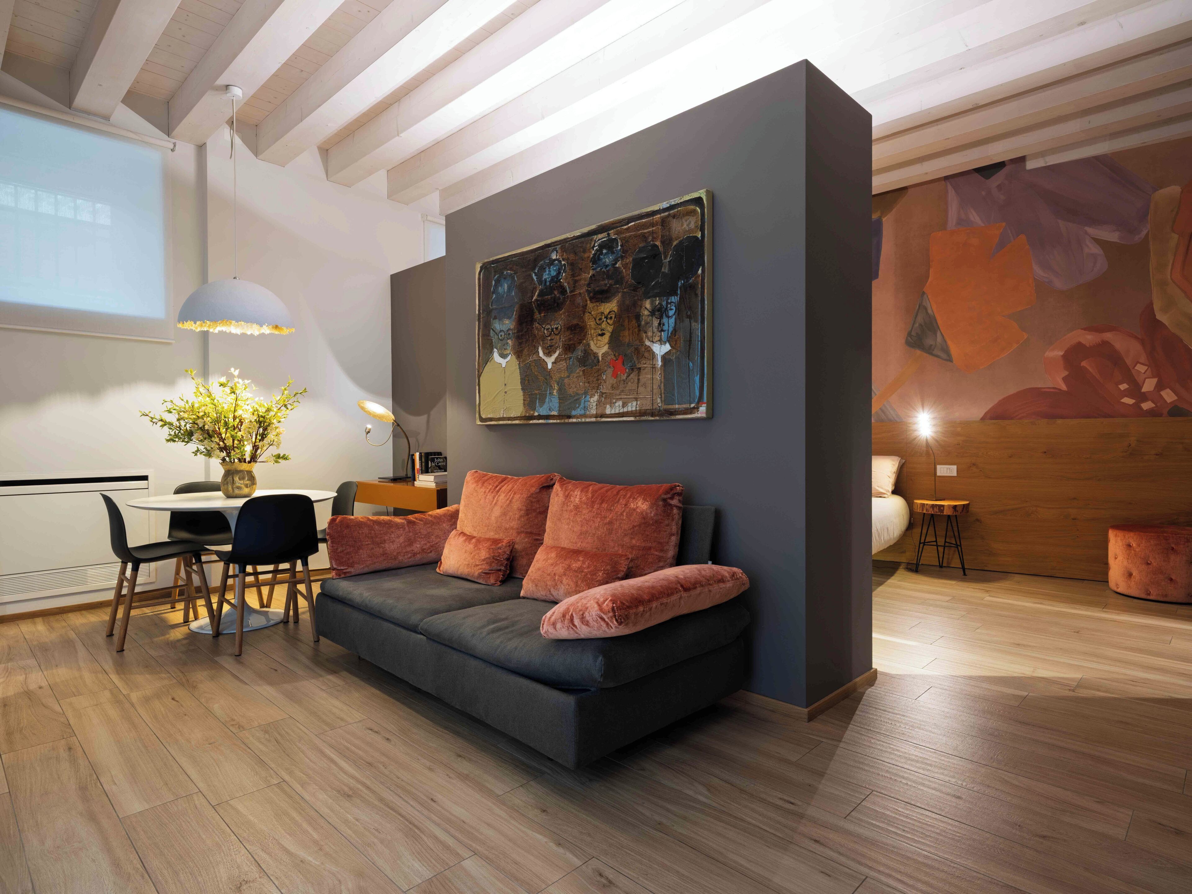 A modern living room with a dark grey sofa, a dining table with chairs, a vase with flowers, and a large abstract painting on the wall.