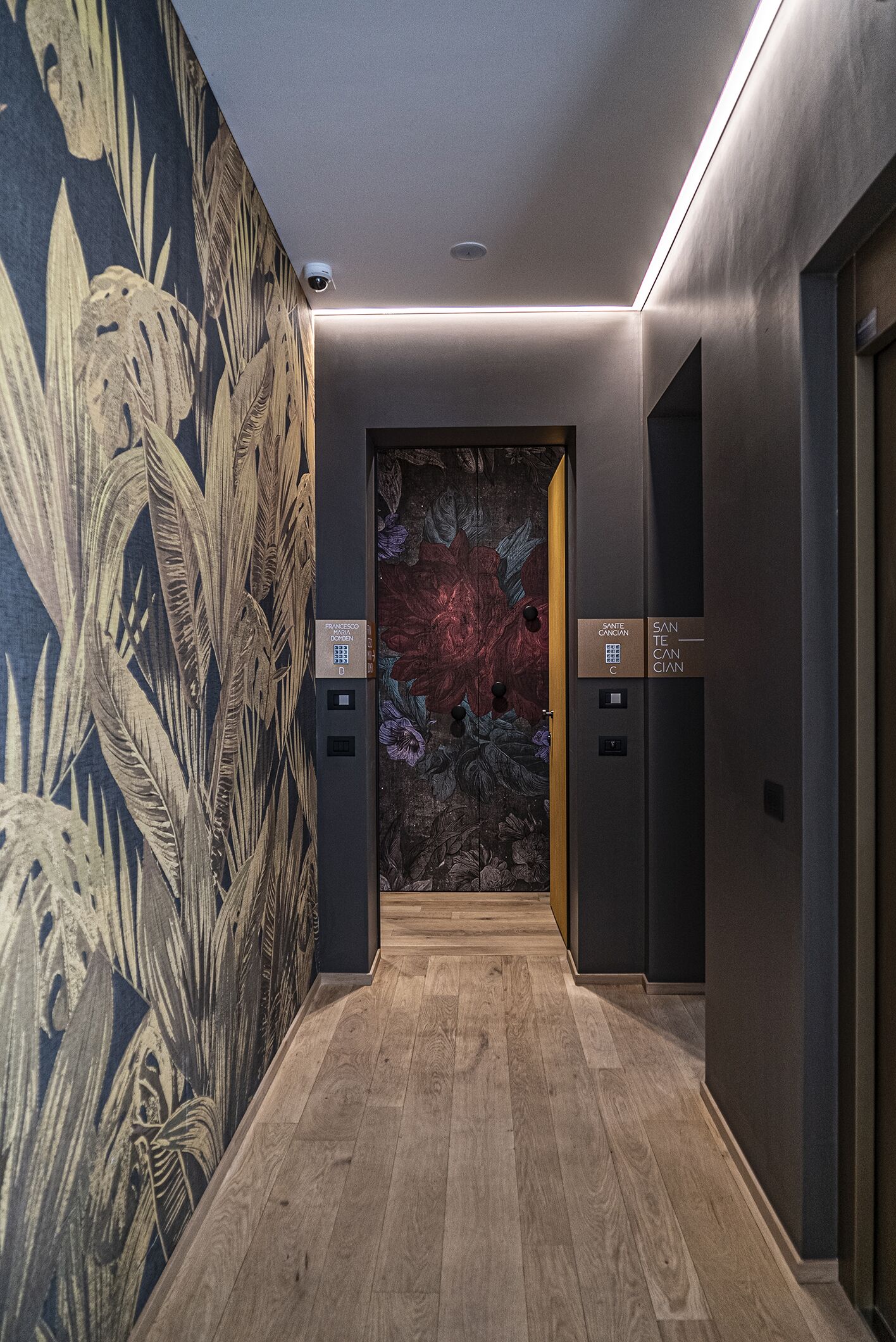 Un ascensore moderno con display digitale e pannello di controllo, circondato da un pavimento in legno e da una parete con motivo a foglia tropicale.