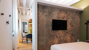 A bedroom with a bed, a TV mounted on the wall, a wooden floor, and a dining area with chairs and a table. - Leoni di Collalto Palace (Treviso)