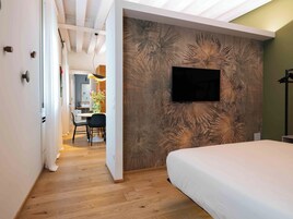 Un dormitorio con cama, televisor montado en la pared, piso de madera y una zona de comedor con sillas y mesa.