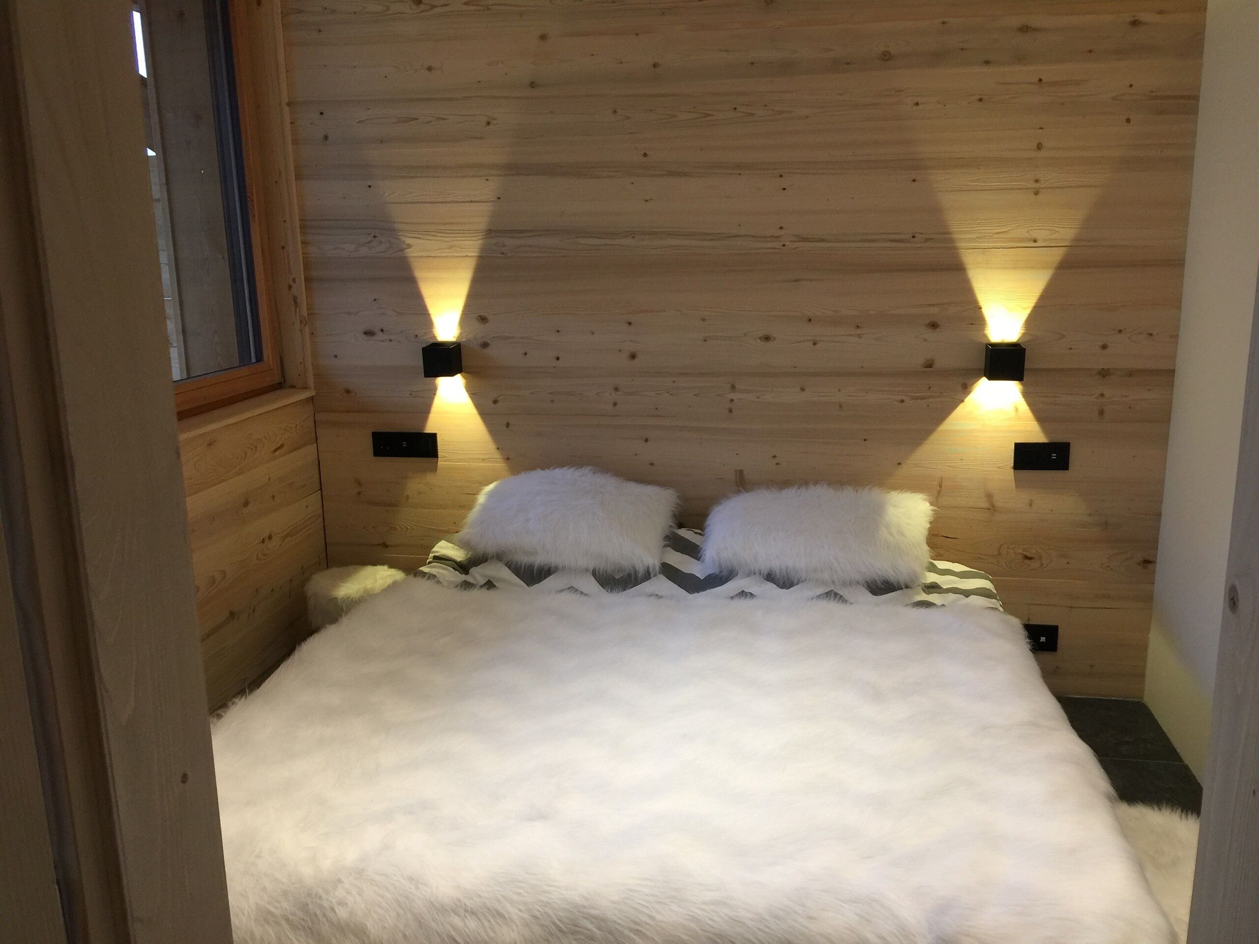 Une pièce chaleureuse avec des murs en bois, un lit, deux lampes fixées au mur et une fenêtre.
