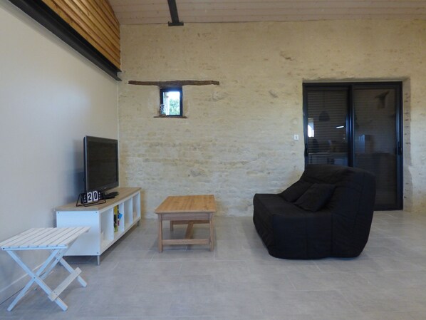 A modern living room with a flat-screen TV on a white stand, a black sofa, a wooden coffee table, and a small shelf with various items.