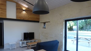 A modern living room with a wooden ceiling, a television on a white stand, a blue sofa, and a large window with a view of an outdoor patio.