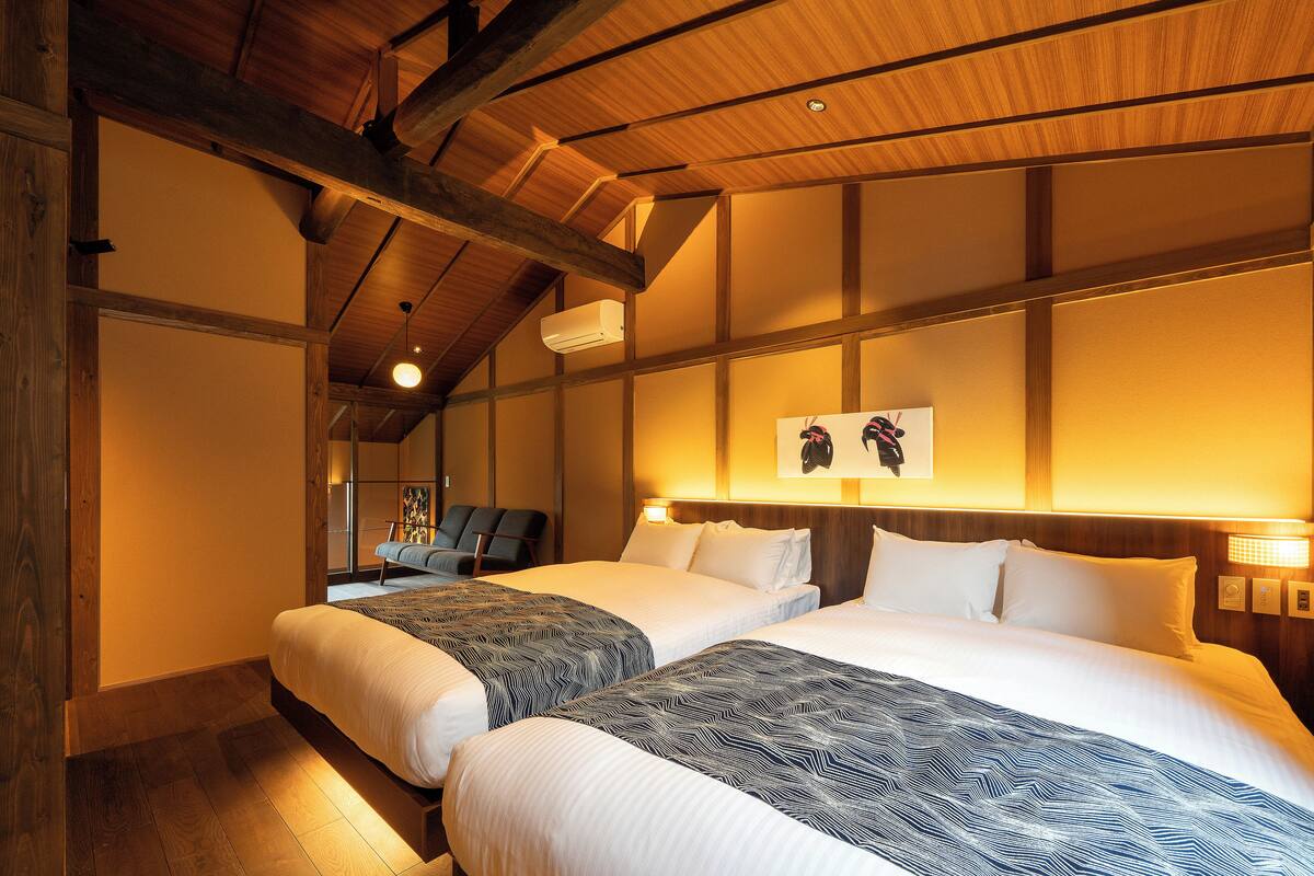 Une chambre d’hôtel avec deux lits, des poutres en bois au plafond, un climatiseur fixé au mur et un tableau encadré au-dessus des lits.