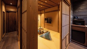 Une pièce de style japonais traditionnel, avec un sol recouvert de tatamis, une petite table basse avec des coussins, un téléviseur et un coin cuisine équipé d’une machine à café.
