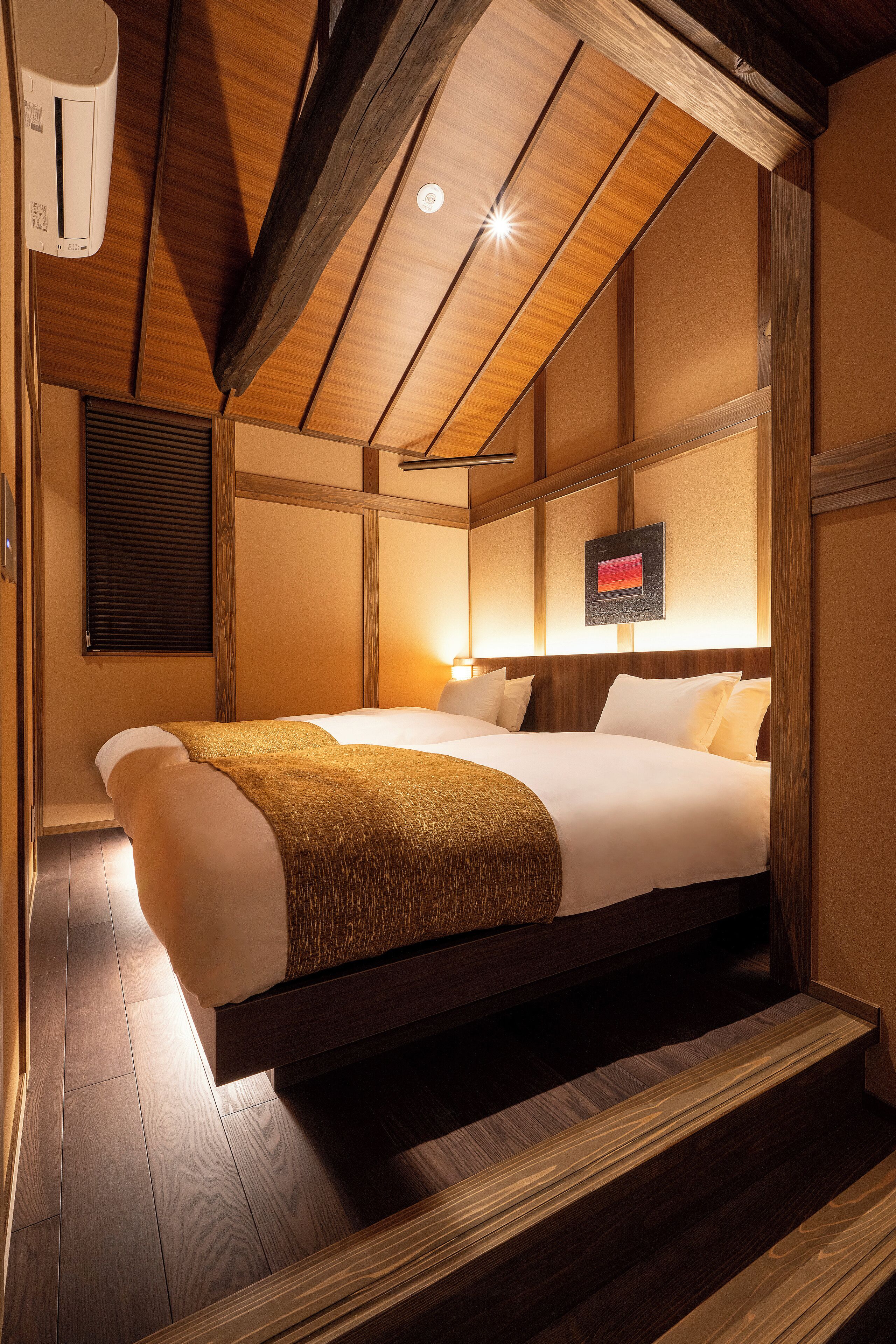 A hotel room with a bed, wooden ceiling beams, a wall-mounted air conditioner, and a framed picture on the wall.