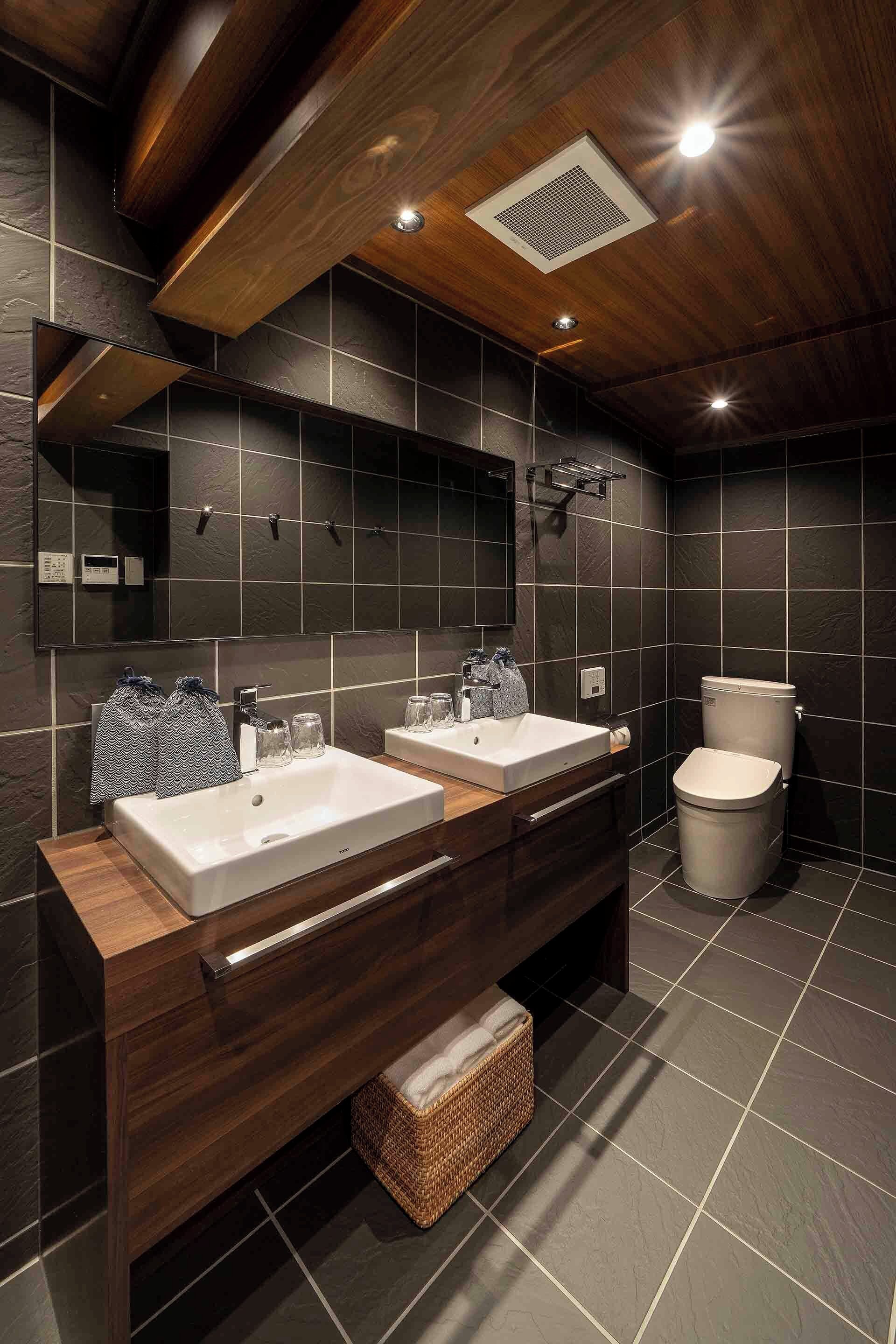 A modern bathroom with dark tiles, wooden vanity, dual sinks, and a toilet.