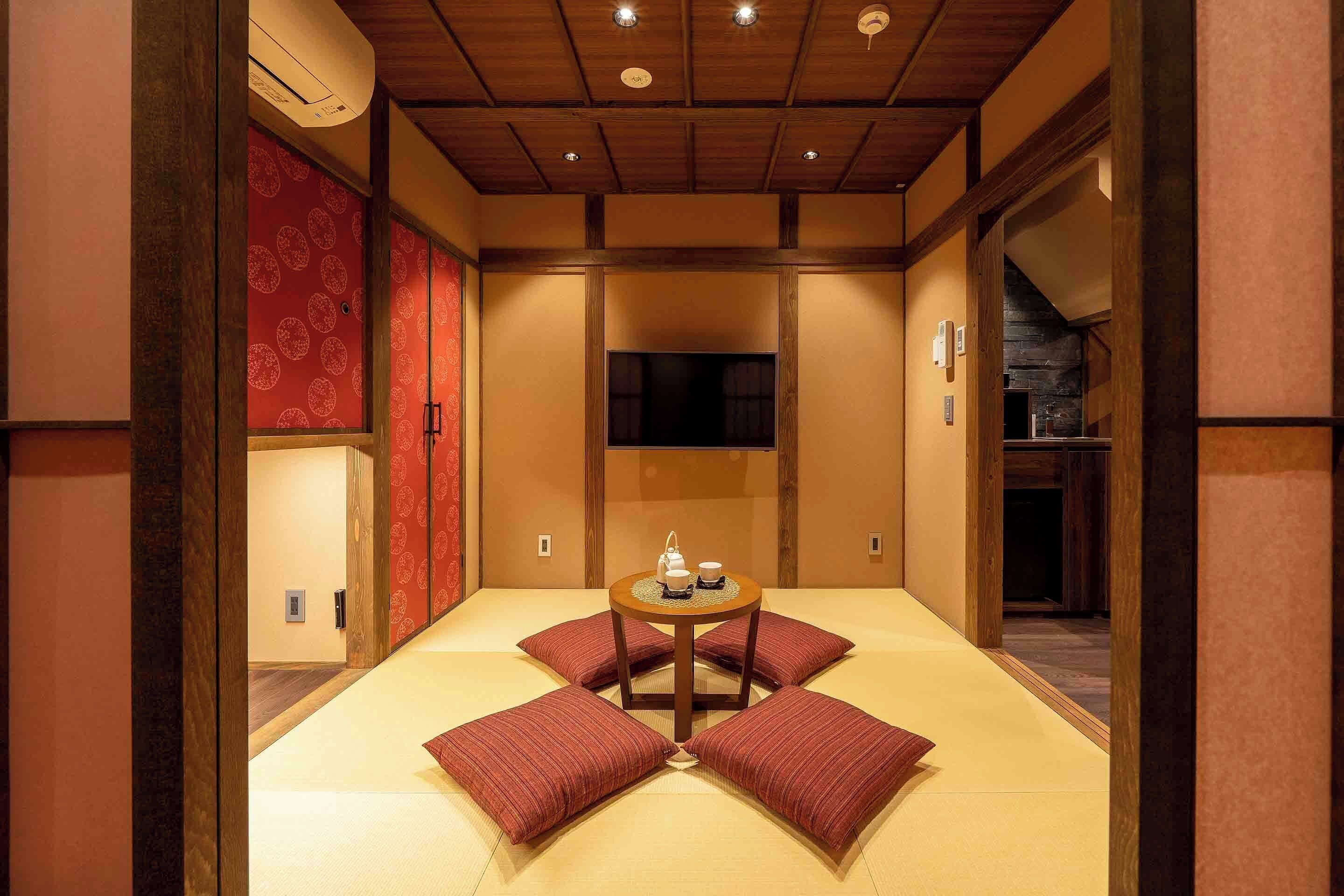 A traditional Japanese room with tatami flooring, a low table with a tea set, and red cushions.
