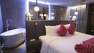 Una habitación de hotel moderna con una bañera grande, una cama con ropa de cama blanca y almohadas rosas, una mesita de noche y un espejo sobre la cama.