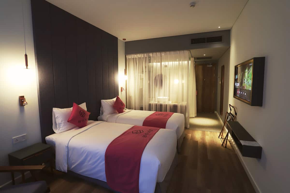 Hotelkamer met twee bedden, elk met een rode deken en een kussen met het logo van het hotel. Er is een bureau met een stoel, een televisie aan de muur en een raam met dunne gordijnen.