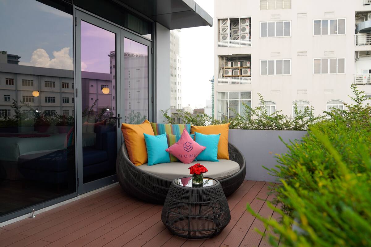 Een balkon met een zithoek met kussens, een glazen tafel met een bloevas, en uitzicht op een stadsgezicht.
