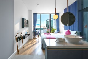 A modern hotel room with a large bed, a round mirror, a bedside table with a white basin, a desk with a chair, and a view of the city through large windows. - HOME Hotel (Ho Chi Minh City)
