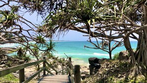 Un sentier en bois mène à une plage aux eaux turquoise et limpides, bordée d’une côte rocheuse.