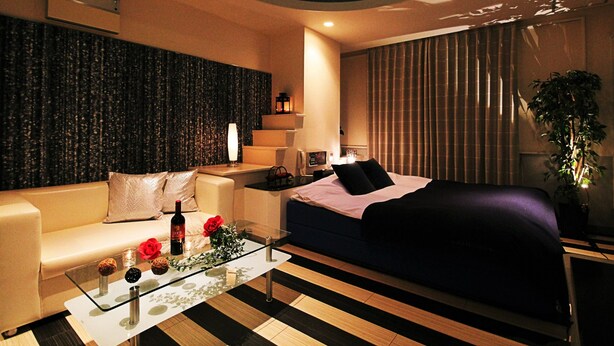Une chambre d’hôtel moderne avec un canapé, un lit recouvert d’une couverture foncée, une table basse en verre agrémentée d’objets décoratifs, un climatiseur fixé au mur et une grande fenêtre avec des rideaux.
