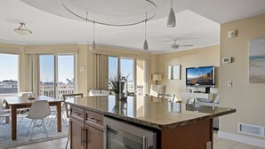 A modern kitchen with a large island, dining area with a table and chairs, and a living area with a sofa and TV. - Gateway Grand 403, Decorator furnished side view unit in a luxury oceanfront building (Ocean City)