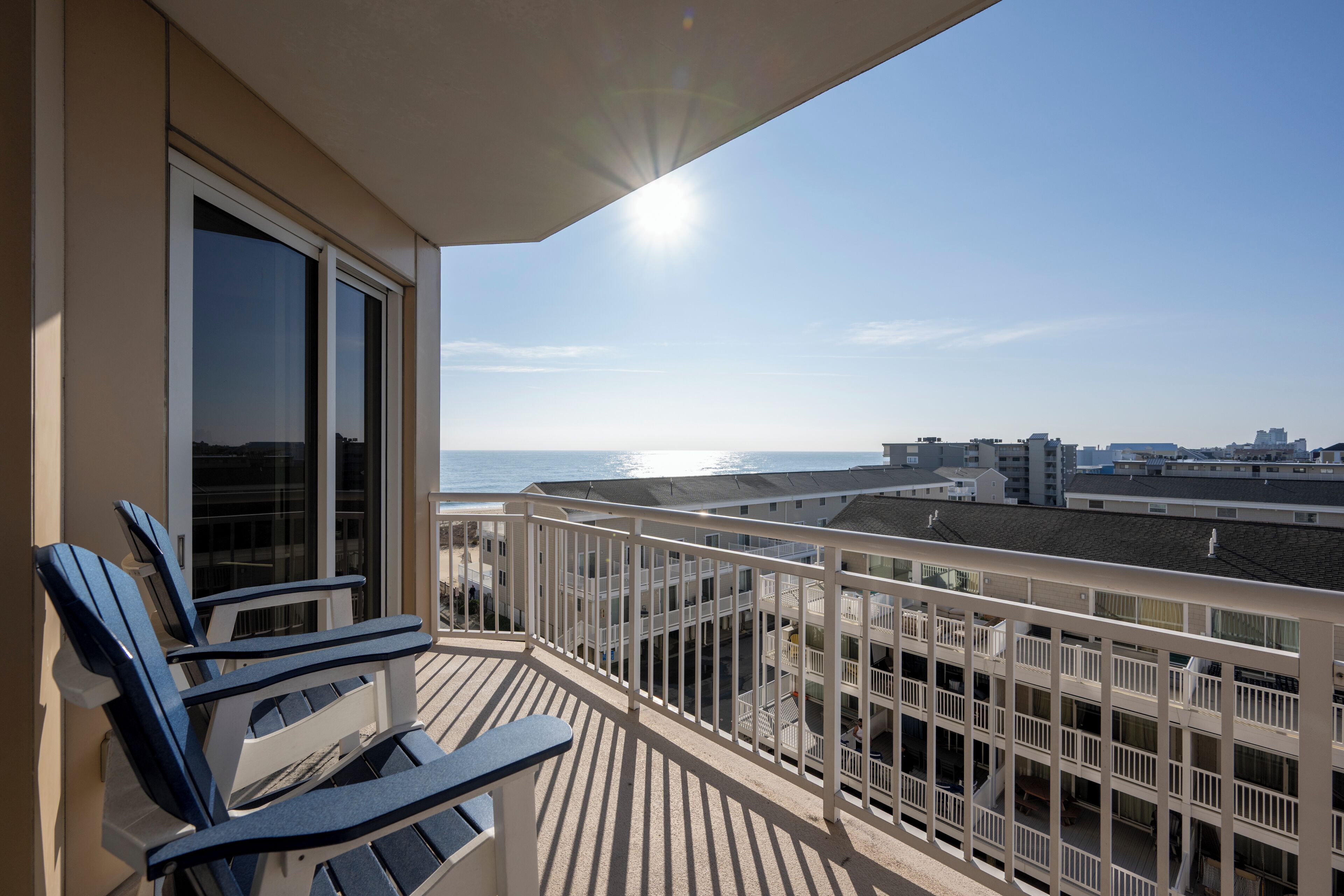 A balcony with two blue chairs, a white railing, and a view of the ocean and buildings.
