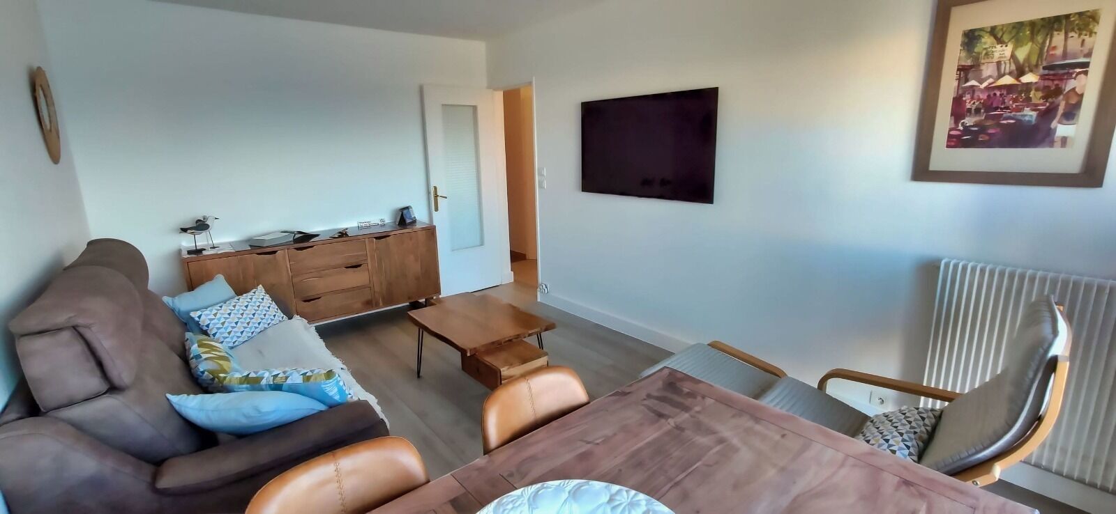 Ein Wohnzimmer mit braunem Sofa, einem Holztisch und einem Esstisch mit Stühlen. Es gibt einen Schrank mit Schubladen und einen flachbildfernseher an der Wand.