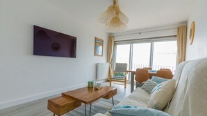 A modern living room with a wooden coffee table, a sofa with patterned cushions, a dining area with a table and chairs, and a large window with curtains.