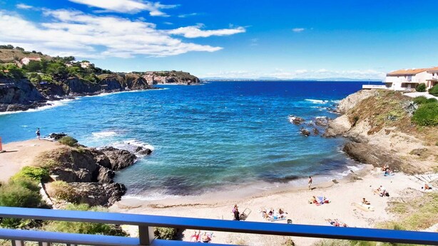 A coastal view with a beach, rocky cliffs, and a row of buildings with balconies overlooking the sea.