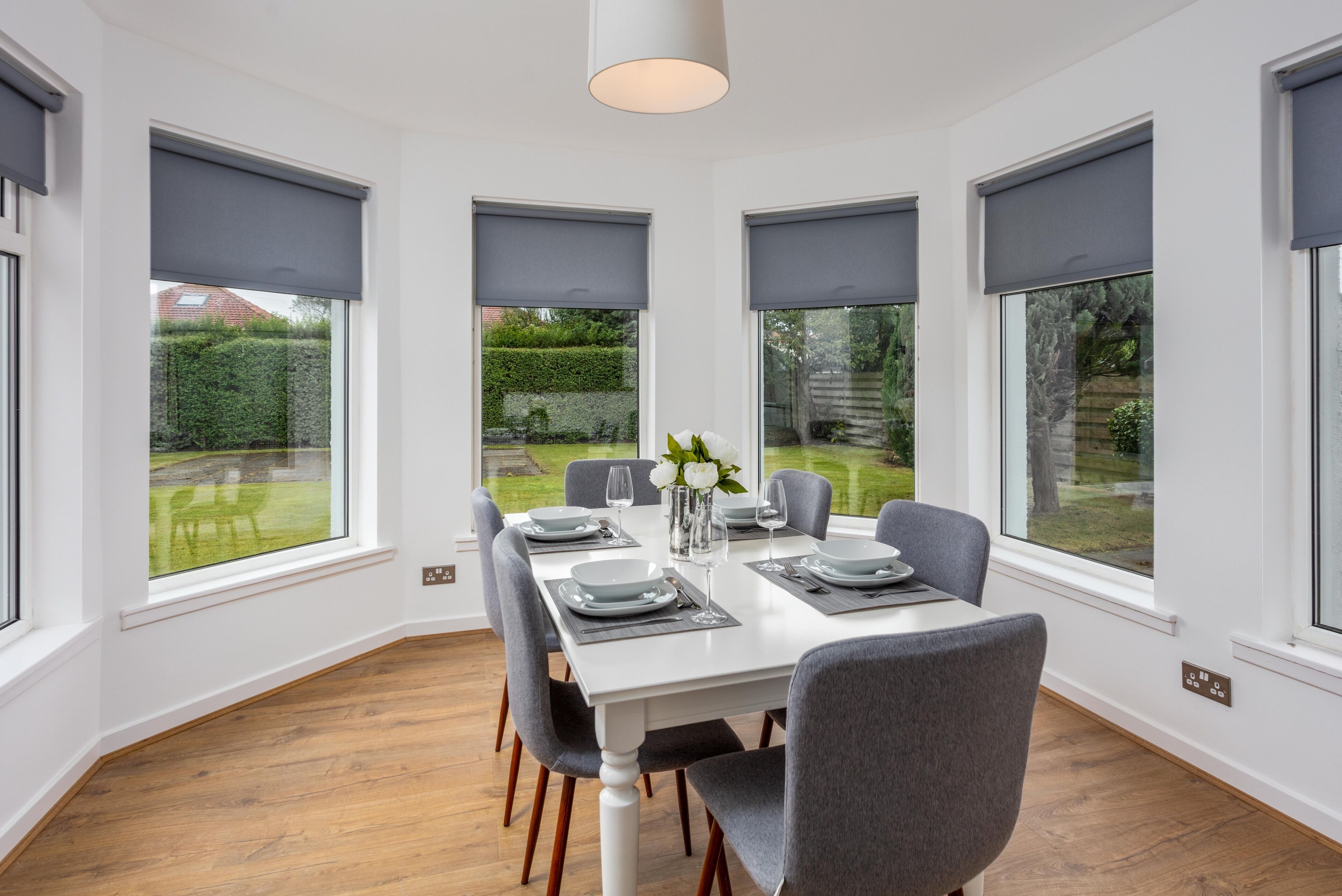 A dining area with a white table set for four, surrounded by grey chairs, large windows with dark blinds, and a view of a garden.
