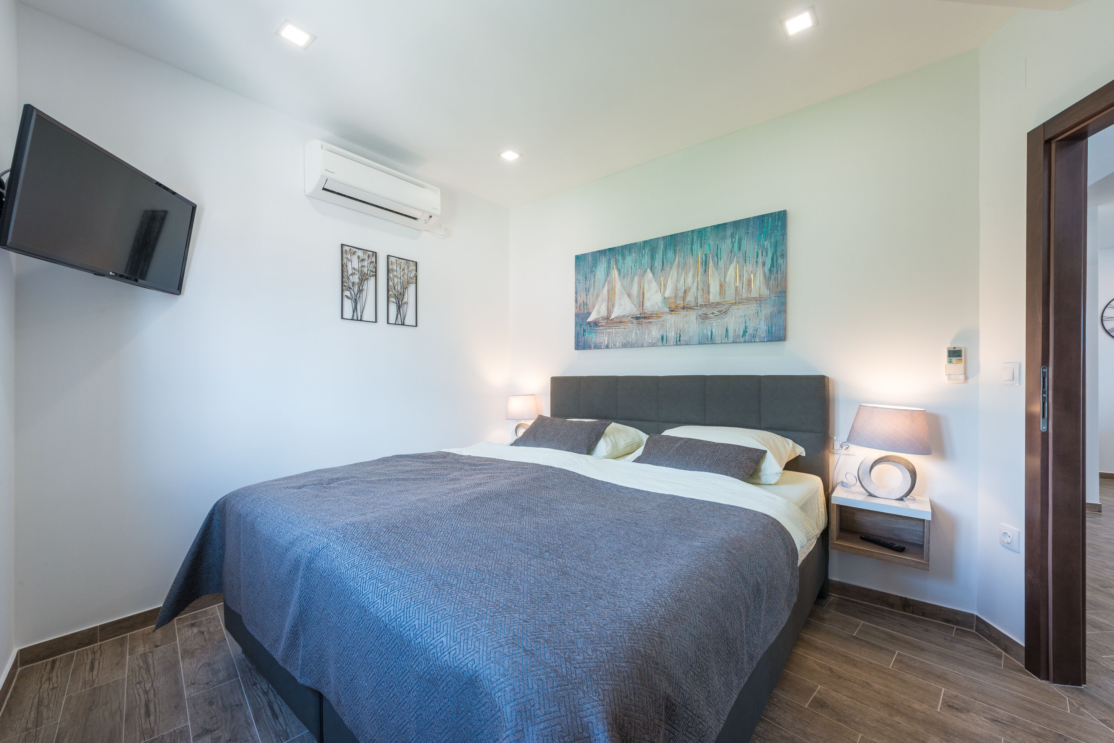 A bedroom with a large bed, a mounted TV, an air conditioner, a painting, and a bedside table with a lamp.