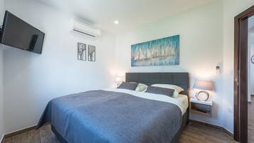 A bedroom with a large bed, a mounted TV, an air conditioner, a painting, and a bedside table with a lamp.