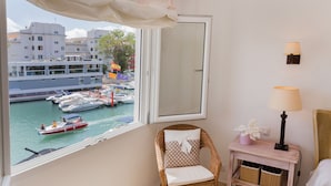 Een kamer met uitzicht op boten in een jachthaven, een rieten stoel met kussen, een kleine tafel met lamp en een rieten mand.