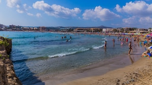 Een strand met helderblauw water, mensen die zwemmen en zonnebaden, en gebouwen op de achtergrond.