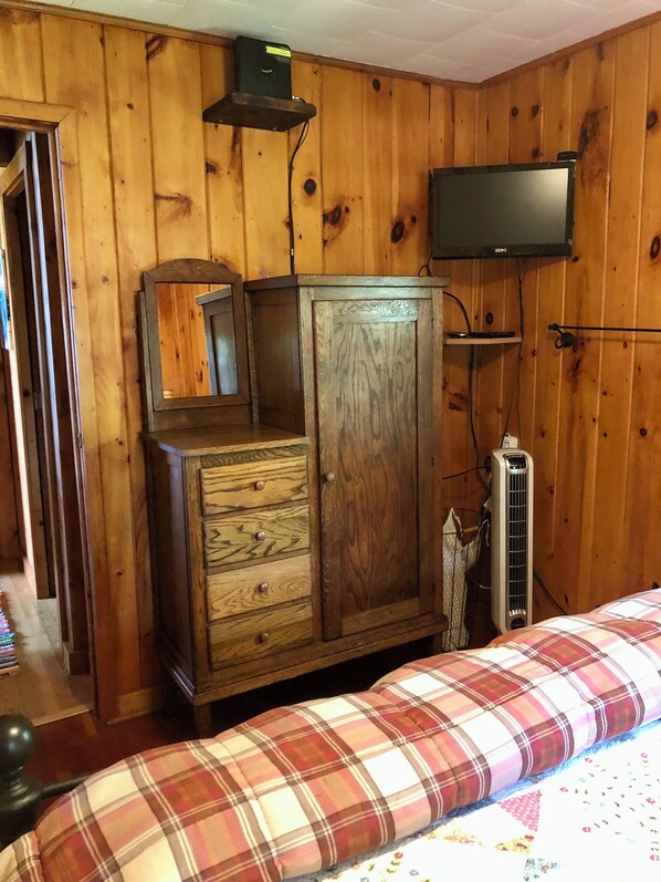 A wooden-paneled room with a bed, a dresser, a television, and a radiator. - Cute As A Bug Cottage On The Shores of Big Manistique Lake (McMillan)